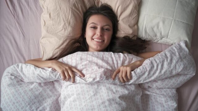Young woman smiling and holding a soft duvet while lying in bed in the morning, embracing comfort and relaxation, enjoying a peaceful moment after waking up