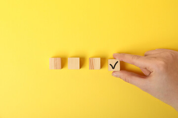 Woman taking wooden cube with check mark on yellow background, top view