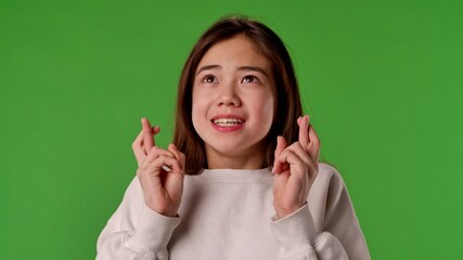 Asian girl child with crossed fingers and please face looking at camera, positive expression, isolated on chroma key green screen studio background mockup.