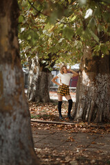 A girl stands on the street among large trees with green leaves. She is wearing a white T-shirt and a short yellow plaid skirt.