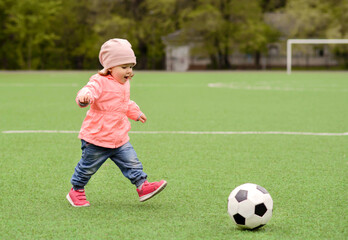Toddler girl playing football: little child in pink jacket, hat, and sneakers running on green soccer field to kick ball. Young sportsman, female power and active childhood concept. Copy space © AMR Studio