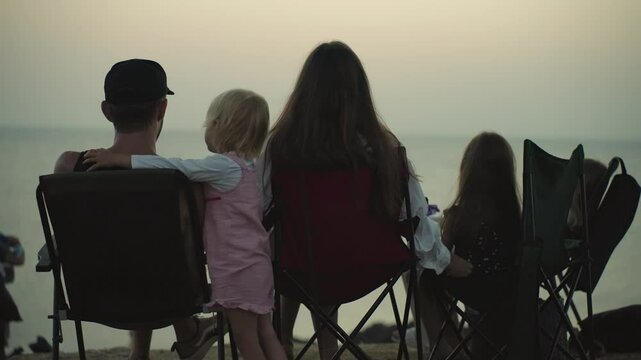Happy family sitting in camping chairs watching the sunset on the sea little daughter hugs and kisses her daddy. Fatherhood Parenthood Togetherness Love Family relationships. Family trip Rest Vacation