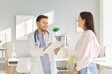Obraz premium Portrait of a young happy man doctor holding report file with appointment in office talking with woman patient and giving consultation during medical examination in clinic. Medicine concept.