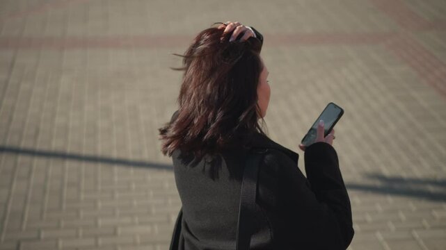 Back view of chic woman in black coat using her phone, touching her hair, ring visible on her finger as she walks on a patterned path, light from the phone screen reflect on her cheek