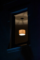 open window of an old house at night showing a dark environment with a chandelier lit in the center of the ceiling that reflects shadows
