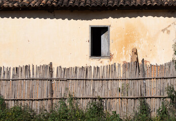 facade of old house with sunburned wall and old clay tiles with a single window half open and half in darkness with small fence made of bamboo pieces on a strong sunny day that produces strong shadows