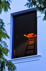 open window of an old house showing a dark environment with a ladder that goes up to the illuminated attic
