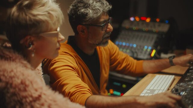 Young stylish female singer and experienced sound engineer working together on new album in recording studio, adjusting audio settings on mixing console and discussing music