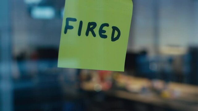 Close-up of a businessman sticking a yellow sticky note with FIRED written on it to a glass surface in an office setting. Concept of job loss, termination, corporate decisions, and unemployment