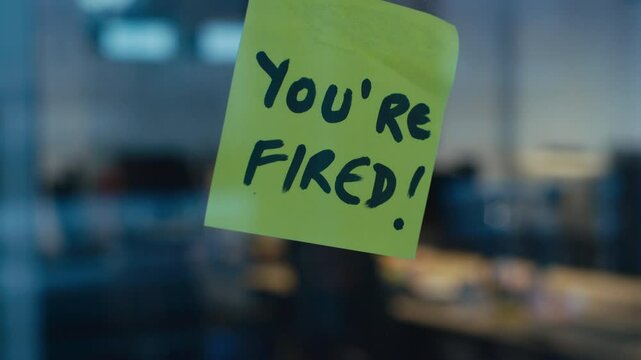 Yellow sticky note with bold black handwriting reading You're fired is attached to a glass surface, with a blurred office environment in the background. Concept of job termination, layoff