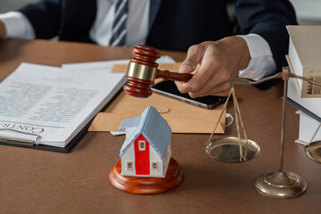 Lawyer striking gavel on desk with model house and scales of justice, reviewing legal documents.