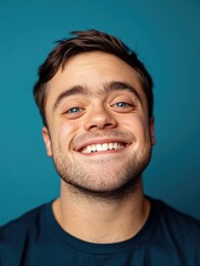 Obraz premium Happy young man with beard smiling and looking directly at the camera, wearing casual attire against blue background.
