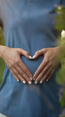 Close-up of a woman forming a heart shape over her stomach with her hands, symbolizing love, affection, and emotional connection in a serene outdoor setting.