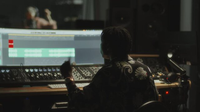 Black music producer sitting at mixing console, working on vocal track displayed on screen, giving thumbs up to stylish female singer performing in studio recording booth. Rack focus
