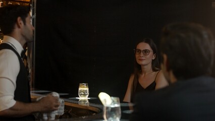 Focusing on beautiful woman guest sitting in special drink gin fizz at luxurious counter bar at vibrant nightlife party atmosphere. Smart bartender wiping glass while talking with customer. Vinosity.