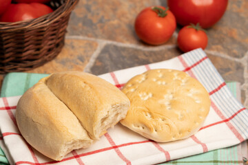 Un par de panes de trigo, una marraqueta y una hallula sobre un paño de cocina, al fondo un canasto con tomates y tomates rojos y maduros