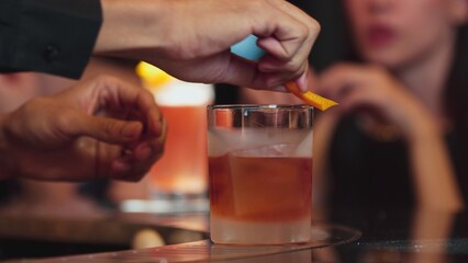 Bartender squeezing orange peel smelling over glass cocktail whiskey putting into Old fashioned on top of rock ice making special drink advertisement alcohol on Paloma and guest background. Vinosity.