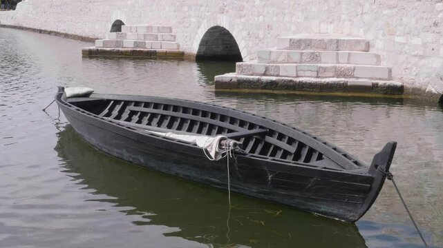 Black wooden fishing boat floating in the water along an old stone bridge.