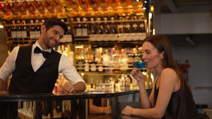 Sitting in smiling beautiful woman guest lively taking with smart bartender while drinking mixed vodka in blue hawaii cocktail and whiskey at luxurious counter bar on Friday meeting at club. Vinosity.