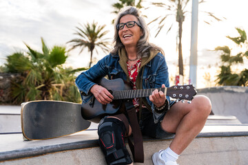 Beautiful active senior woman playing guitar and singing - Mature adult female skateboarder having fun, concepts about elderly, modern lifestyle and green mobility