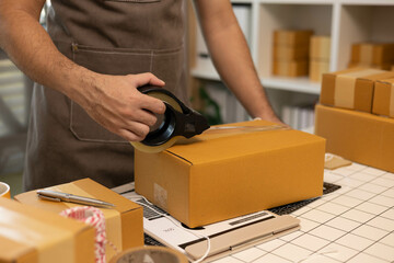 A man is sealing a brown cardboard box with tape. The box is on a table with a laptop and a stack...