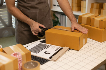 A man is sealing a brown cardboard box with tape. The box is on a table with a laptop and a stack of papers. The man is wearing an apron and he is in a focused and determined mood
