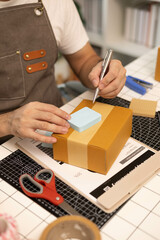 A man is writing on a cardboard box with a marker. He is wearing an apron and has scissors on the table
