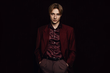 handsome blonde young guy stands with his hands in his pockets and a neutral face expression, looking at the camera in a strong and confident way, exuding poise and self-assurance