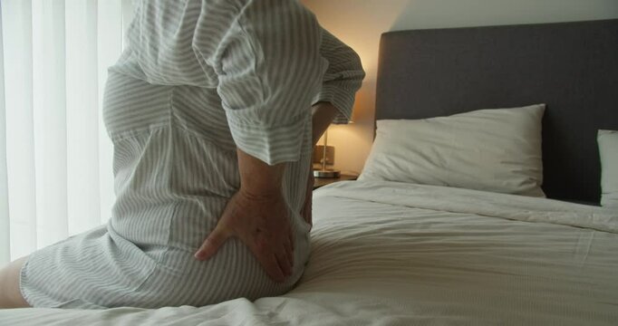 An elderly woman experiences back pain while sitting on her bed in a cozy bedroom and holding her lower back with both hands to relieve the pain