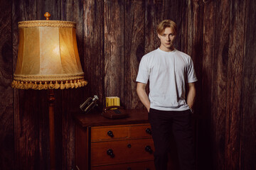 hot guy stands in an old retro living room, wearing a white shirt with his hands in his pockets of pants, leaning on a sideboard and looking at the camera with a confident, bored, and sexy expression