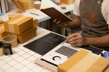A man is writing in a notebook while holding a cardboard box. He is wearing an apron and he is working in a warehouse
