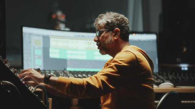 Senior music producer adjusting controls on mixing console and enjoying sound as working at digital workstation, singer performing in recording booth in background