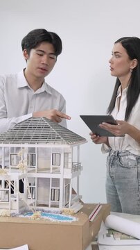 Beautiful young caucasian project manager brief asian engineer by using tablet while engineer measuring house model on meeting table with blueprint, architectural equipment scatter around. Immaculate