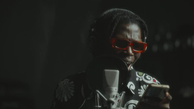 Black male musician wearing headphones and orange sunglasses, reading lyrics from phone and singing into microphone in recording studio booth