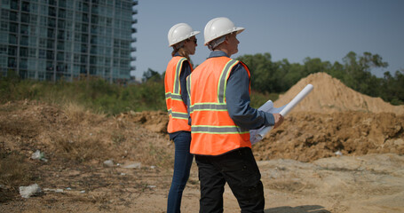 The project engineer team with orange safety vests  holds laptops and blueprint drawings working at the construction sideline, drainage construction