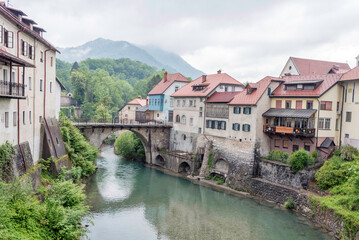 Scenic view of &Scaron;kofja Loka, Slovenia, in spring. A historic stone bridge and medieval houses reflect in the calm river, surrounded by lush greenery and mountains. Charming European village atmosphere.