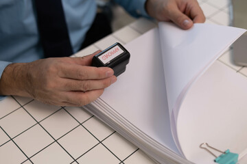 A man is sitting at a desk with a stack of papers and a pen. He is holding a stamp and he is in the process of stamping something. The scene suggests a professional or business setting
