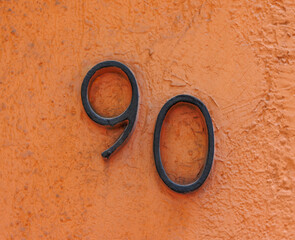 Black metal street address number, number 90, mounted on an orange stucco wall.