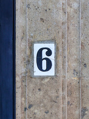 Street address number, number six (6). Cracked white tile with a dark blue number inset into a brown carved stone wall.