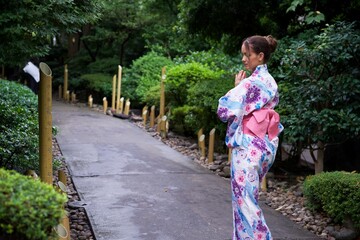 A woman wearing a floral kimono stands in meditation on a peaceful garden pathway, surrounded by lush greenery and decorative bamboo lanterns. 