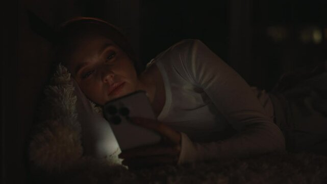 Young woman lying in bed, using smartphone at night, illuminated by soft glow of the screen, battling insomnia scrolling through late-night social media, turns off phone and going to bed, close-up.