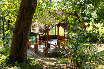 Cozy wooden gazebo with brick columns in lush garden surrounded by tall green trees and foliage.
