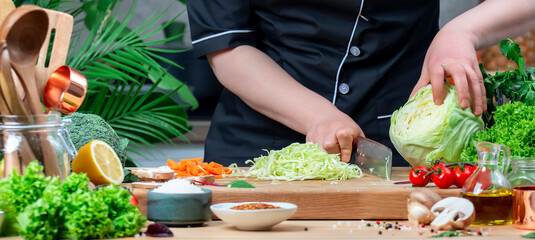 Chef pushes aside chopped cabbage with knife. Cozy kitchen with wooden table, kitchenware, vegetables, herbs and ingredients for cooking. Healthy vegan eating, culinary, recipes, food blogging