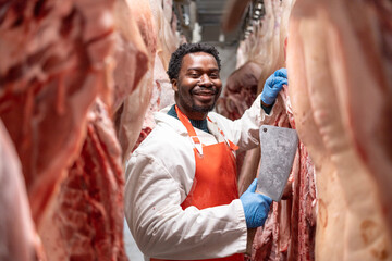 Portrait of smiling African American butcher standing by pork meat hanging inside slaughterhouse.
