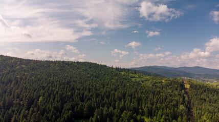 Naklejka premium Majestic Mountains, Lush Green Forests, And Soft White Clouds On A Sunny Day. Aerial View Showcasing The Stunning Beauty Of Nature From A High Perspective. 