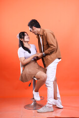 a couple of young lovers happy expression where the man is standing and the woman is sitting on a white chair, face to face very romantic. isolated on orange background