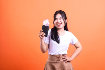 Happy woman holding a wallet filled with cash on an orange background posing with hand on hip