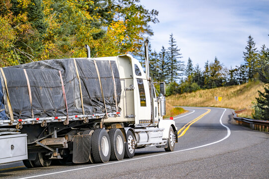 Industrial standard big rig white semi truck tractor with extended cab transporting covered and fastened cargo on flat bed semi trailer running on the winding mountain road with autumn trees