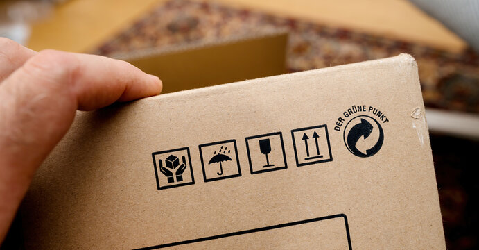 A person holds a Hi-Fi equipment package made of cardboard, featuring handling symbols for fragile, keep dry, and proper orientation, along with the Der Grune Punkt recycling mark.