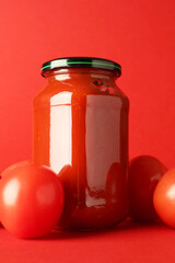 Ketchup in glass jar and fresh tomatoes on red background, closeup